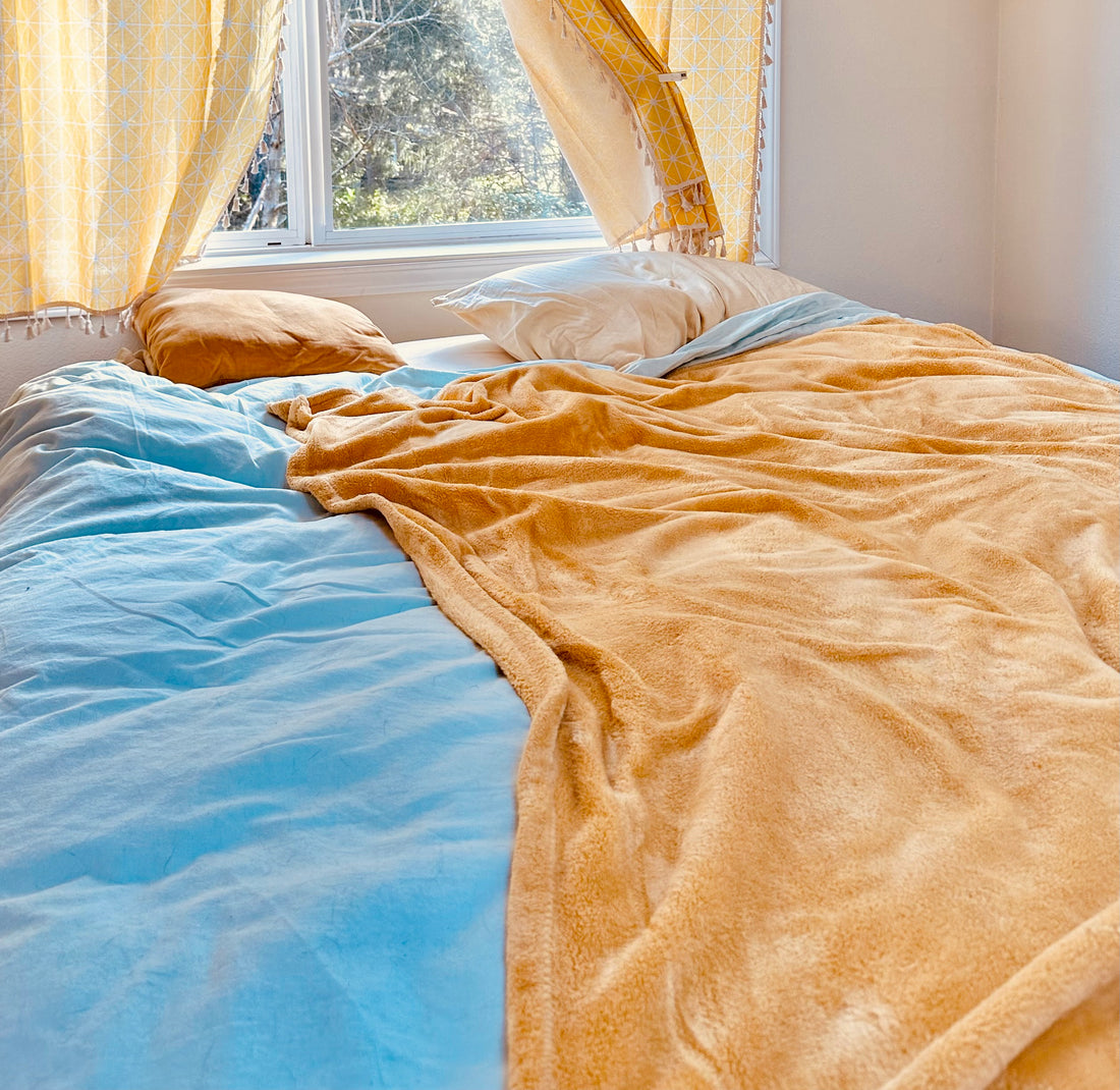Image of a bed with yellow and blue blankets, the sun shining inside from a window above.